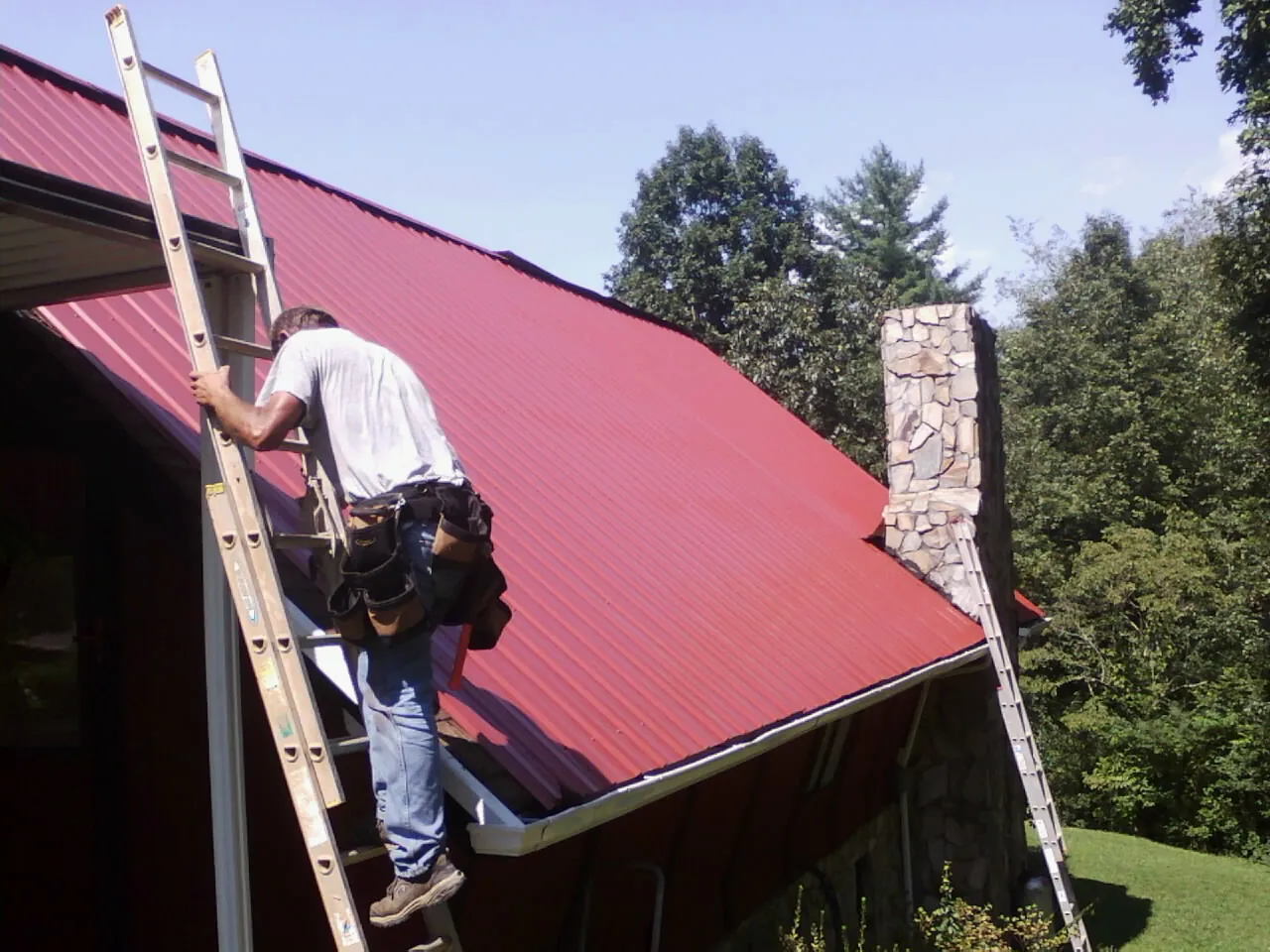 Roofer on a ladder beside a red metal roof and stone chimney — Soffit & Fascia Repair by Roofing Brothers in Union Grove
